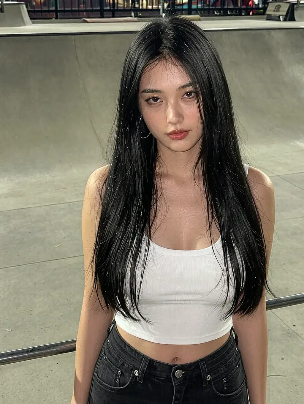 Long straight black hair, white crop top and black jeans, small hoop earring, at a concrete skatepark